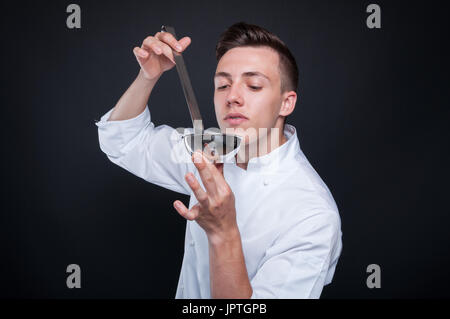 Erfolgreiche männliche Koch Verkostung seine Suppe aus Edelstahl Pfanne auf dunklem Hintergrund isoliert Stockfoto