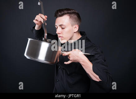 Junge männliche Koch mit großen Topf riecht das Essen aus der Pfanne auf dunklem Hintergrund Stockfoto