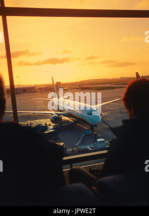 Passagiere warten in einer vollen Abflughalle neben Fenstern mit einem Ryanair Boeing 737-800 in der Abenddämmerung vor geparkt. Stockfoto