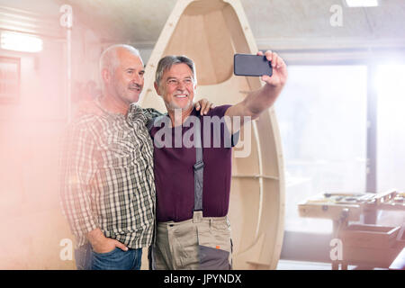 Stolz, lächelnd männlichen Tischler mit Kamera-Handy nehmen Selfie nächsten zu Holz-Boot in Werkstatt Stockfoto