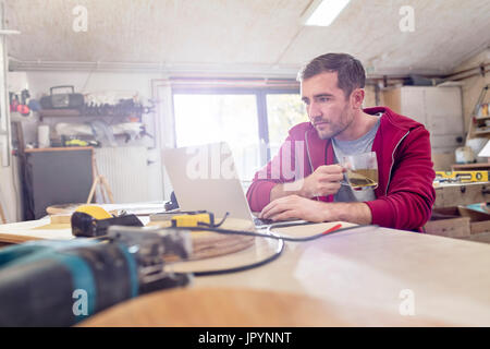 Männliche Zimmermann, Tee trinken und arbeiten am Laptop auf Werkbank in Werkstatt Stockfoto