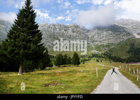 Junge in der Mitte von einem Wanderweg mit herrlichem Bergblick auf das Hochland von Montasio, Julische Alpen, Friaul Julisch Venetien, Italien springen. Stockfoto