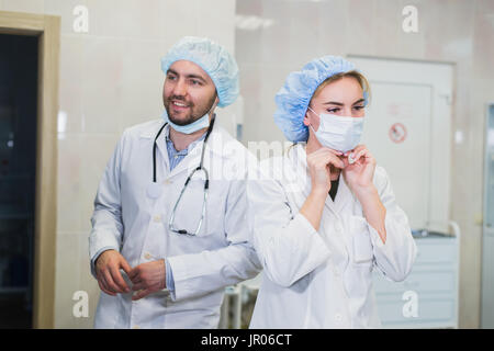 Zuversichtlich, Ärztin, auf medizinische Gesichtsmaske während der Vorbereitung für Betrieb, ihr männlicher Kollege hinter ihr stehen Stockfoto