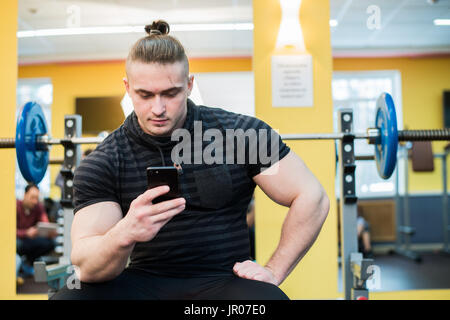 Hübscher Kerl SMS auf seinem Smartphone im Fitnessstudio. Stockfoto