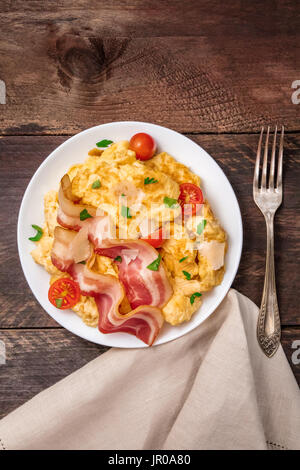 Eine Overhead Foto von einen Teller mit Rührei mit Speck, Tomaten, frische Petersilie und geriebener Käse, geschossen von oben auf eine dunkle rustikale Hinterg Stockfoto