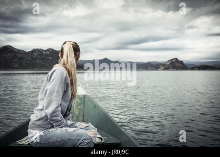 Nachdenkliche junge Frau Touristen in schöner Landschaft, am Bug des Bootes auf dem Wasser schwimmend in Richtung Ufer im bewölkten Tag mit dramatischen Himmel Stockfoto