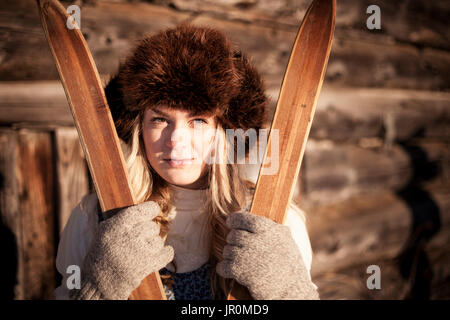 Portrait eines Mädchen mit einem Pelzhut und halten ein paar Holz- Skier an ein Blockhaus; Homer, Alaska, Vereinigte Staaten von Amerika Stockfoto