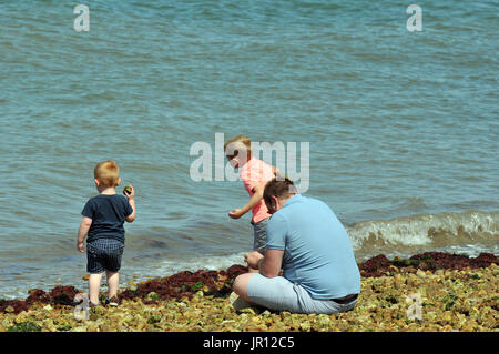 Ein junger Mann sttig auf einem steinigen Strand auf der Suche nach seiner Söhne oder zwei kleine Jungen werfen und skimming Steine im Meer am Meer. Stockfoto