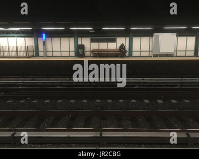 Mann in Hoodie sitzt auf Holzbank Blick auf sein Handy warten auf die u-Bahn in der Nacht. Spät in die Nacht warten auf die New Yorker u-Bahn zu trainieren. Stockfoto