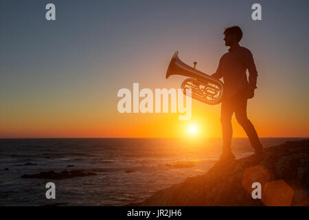 Silhouette der Musiker mit Trompete auf felsigen Küste während des Sonnenuntergangs. Tuba. Stockfoto