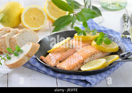 Zwei gebratene Lachsfilets mit gedünstetem Zitronenscheiben in einer Pfanne, serviert mit Baguette, weißen Wein im Hintergrund Stockfoto