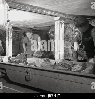 1948, historische England, ein Bergmann mit einem Spaten große Klumpen von Kohle und anderen mineralischen Materie in einer Metal Band unter dem wachsamen Auge eines erfahrenen Bergbau Supervisor zu laden. Stockfoto