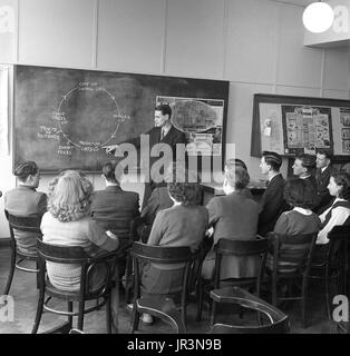 1948, historische, post-ww2 Großbritannien, eonomics Lektion, eine männliche Lehrer mit einem Diagramm auf einer Tafel zu einer Gruppe von männlichen und weiblichen Studierenden zu erklären, wie die lebenshaltungskosten von Großbritannien benötigen Rohstoffe zu importieren betroffen ist. Stockfoto