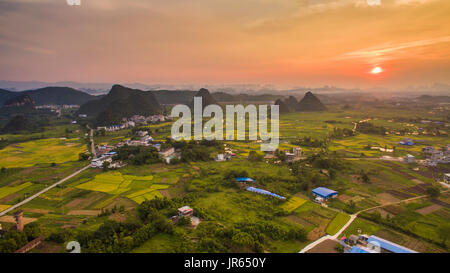 Luftaufnahme eines Dorfes von padi Felder und Hügel gegen die untergehende Sonne in Guanxi, China umgeben Stockfoto