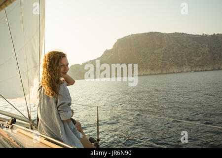 Horizontalen Schuss der jungen blonden Frau genießen Segeln mit Meer und Bergen als Hintergrund Stockfoto
