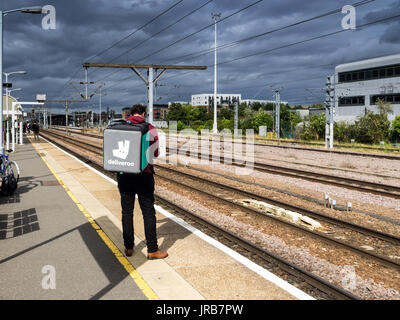 Mit dem Zug - eine Deliveroo Deliveroo fast food Kurier Fahrer wartet ein bummelzug in Cambridge, Großbritannien Stockfoto