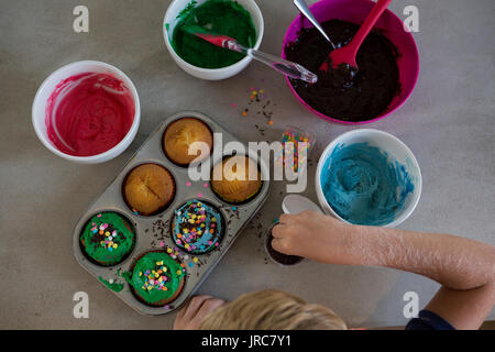 7/8 Hand des jungen dekorieren Cupcakes mit Sprinklern an den Küchentisch Stockfoto