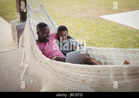 Vater und Sohn mit Laptop beim Entspannen auf einer Hängematte im Garten Stockfoto