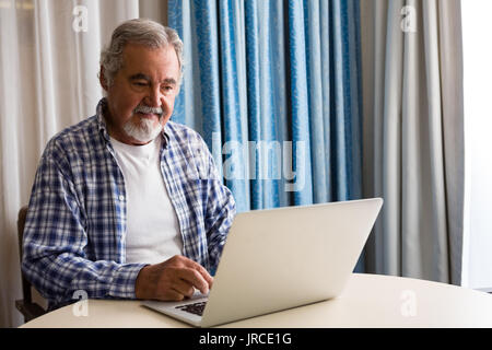 Älterer Mann mit Laptop, während bei Tisch und in der Krankenpflege zu Hause sitzen Stockfoto