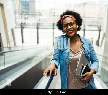 Junge afrikanische Frauen Unternehmer selbstbewusst lächelnd, während sie Reiten eine Rolltreppe in der Lobby eines modernen Bürogebäudes, einen Laptop und Verschleiß Stockfoto