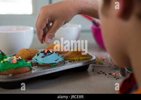 7/8 Hand des jungen dekorieren Cupcakes mit Sprinklern an den Küchentisch Stockfoto