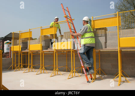 Ein Maurer klettert eine Leiter auf eine vollständig geschützte Arbeitsbühne. Ein anderer baut eine Wand aus thermischen Betonblöcken. Wohnsiedlung in Großbritannien. Stockfoto