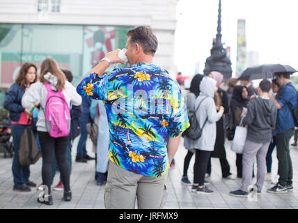 Ein Mann trägt ein Hawaian T-shirt beim Menschen Unterschlupf unter Sonnenschirmen aus einem Regenschauer an der Southbank, London, da das nasse Sommerwetter weiter. Stockfoto