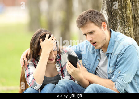 Traurige Frau zeigt negative Nachricht an einem überraschten Freund saß auf dem Gras in einem Park Stockfoto