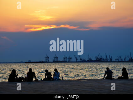 Silhouetten von Menschen, die am Strand vor dem Hafen von Thessaloniki Griechenland sitzen, den Sonnenuntergang zu beobachten. Stockfoto