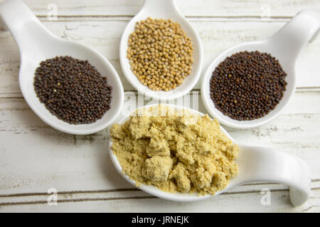 In der Nähe von trockenen Senfpulver und ganze Senfkörner in weißes Porzellan Löffel auf rustikale Holzplanken. Stockfoto