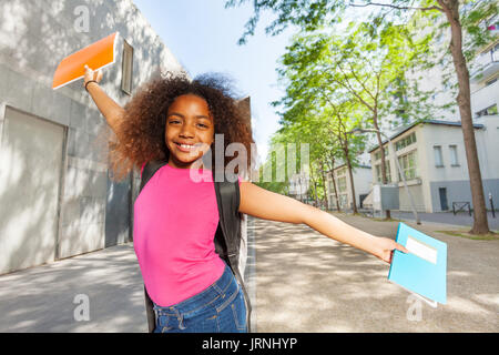 Lächelnd Closeup Portrait von curly Afrikanische Mädchen, dass Lehrbücher und die Verbreitung von Händen in der Nähe der Schule Stockfoto