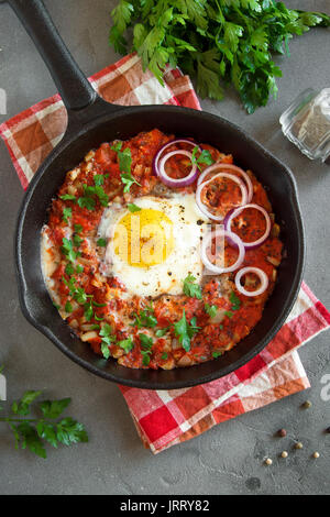 Shakshuka in einer gusseisernen Pfanne. Mitten im orientalischen Stil Eier in tomaten und paprika sauce mit Petersilie, Zwiebeln und Gewürzen auf grauem Beton Hintergrund pochiert. Stockfoto