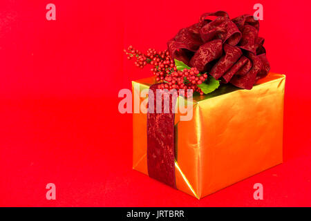 Ein Geschenk in Gold Folie umwickelt ist mit einem dunklen Rot, Damast und Bogen Bänder und roten Beeren Dekoration gekrönt.