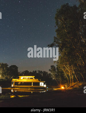 Hausboot auf dem mächtigen Murray River bei Nacht unter den Sternen mit einem Lagerfeuer am Ufer in der Nähe von Echuca. Stockfoto