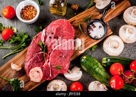 Rohes Fleisch mit Zutaten für das Abendessen. Rinderfilet, Filet, auf ein Schneidbrett, mit Salz, Pfeffer, Petersilie, Rosmarin, Öl, Knoblauch, Tomaten, Pilze, Stockfoto