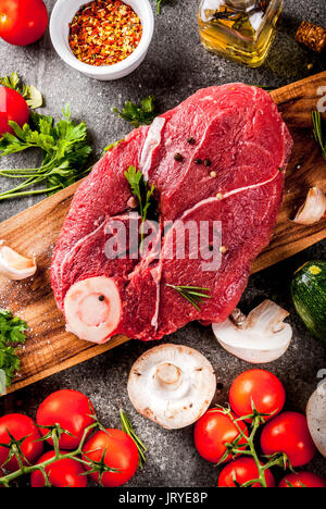 Rohes Fleisch mit Zutaten für das Abendessen. Rinderfilet, Filet, auf ein Schneidbrett, mit Salz, Pfeffer, Petersilie, Rosmarin, Öl, Knoblauch, Tomaten, Pilze, Stockfoto