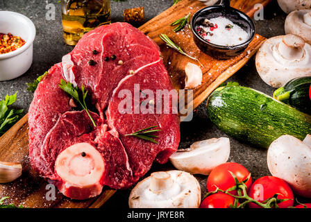 Rohes Fleisch mit Zutaten für das Abendessen. Rinderfilet, Filet, auf ein Schneidbrett, mit Salz, Pfeffer, Petersilie, Rosmarin, Öl, Knoblauch, Tomaten, Pilze, Stockfoto