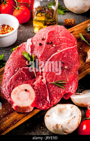 Rohes Fleisch mit Zutaten für das Abendessen. Rinderfilet, Filet, auf ein Schneidbrett, mit Salz, Pfeffer, Petersilie, Rosmarin, Öl, Knoblauch, Tomaten, Pilze, Stockfoto