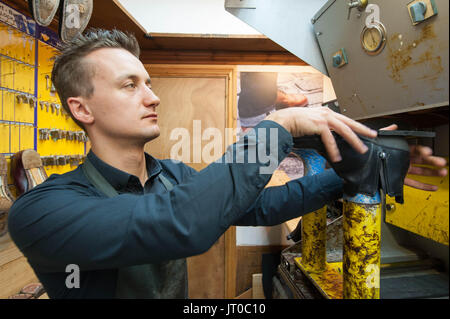 Schuster repariert Lederschuhe in einer traditionellen Werkstatt. Werkzeuge, Schlüsselschneidegeräte und Ersatzsohlen sind an der Wand sichtbar Stockfoto