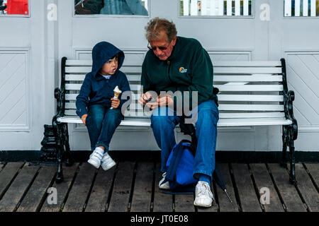 Ein älterer Mann und ein kleiner Junge auf einer Bank sitzen Unterschlupf vor dem Regen am Pier von Brighton, Brighton, Sussex, UK Stockfoto