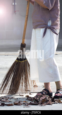 Alte Frau fegt trockene Blatt im Freien auf der Estrich durch die großen langen Besen. Stockfoto