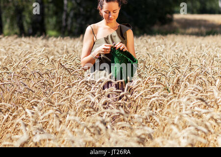 Eine junge Frau, die in einem Sommer top und rock liegt in einem schönen goldenen Feld an einem Sommertag aus Weizen und strickt einen grünen Pullover aus natürlichen Kammgarn t Stockfoto