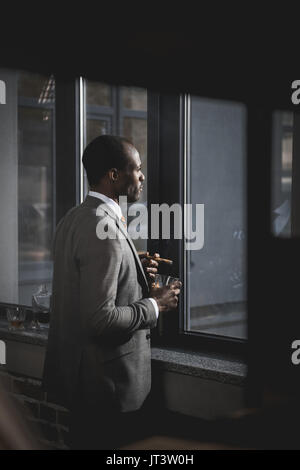 Nachdenklich afrikanische amerikanische Geschäftsmann in Anzug mit Glas Whiskey rauchen Zigarre Stockfoto