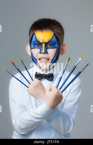 Kleiner Junge mit Gemälden auf seinem Gesicht Wolverine mit Pinsel in der Hand Stockfoto