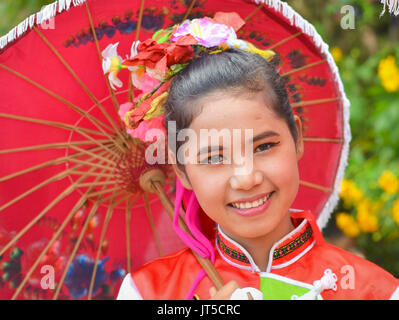 Hübsches thailändisches chinesisches Mädchen im traditionellen Kleid mit rotem Sonnenschirm lächelt für die Kamera. Stockfoto