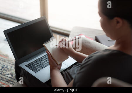 Frau Freelancer schreiben Memo auf kleinen Notizblock während der Arbeit mit dem Laptop. on-line-Geschäft und Selbständige Konzept Stockfoto