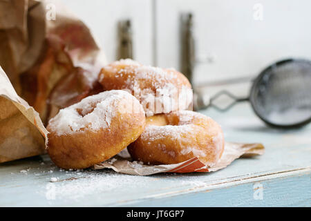 Hausgemachten Krapfen mit Zucker Pulver aus Paper Bag mit Vintage Sieb auf Blau Holztisch serviert. Rustikaler Stil, Tageslicht. Stockfoto