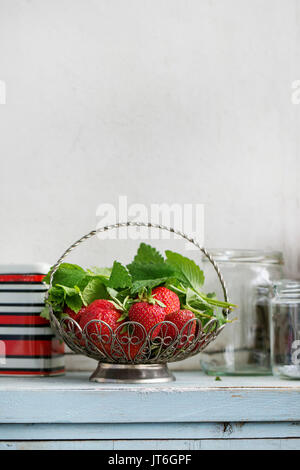 Frische reife Garten Erdbeeren und Melisse Kräuter in vintage Vase stehen mit leeren Glas und Metall Gläser für Stau auf Blau-weiße Küche aus Holz Tisch. Stockfoto