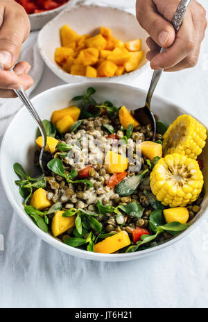 Eine Frau rühren eine Schüssel mit Linsensalat mit Kräutern, Mais und Pfirsiche mit einem Löffel und einer Gabel auf einem weißen Hintergrund. Stockfoto