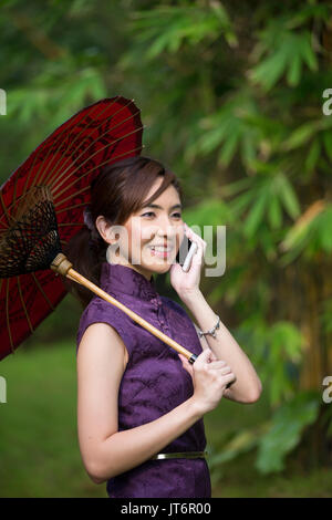 Chinesische Frau sprechen über ein Smart Phone und das Tragen eines Cheongsam chinesische Kleid mit traditionellen Regenschirm. Stockfoto